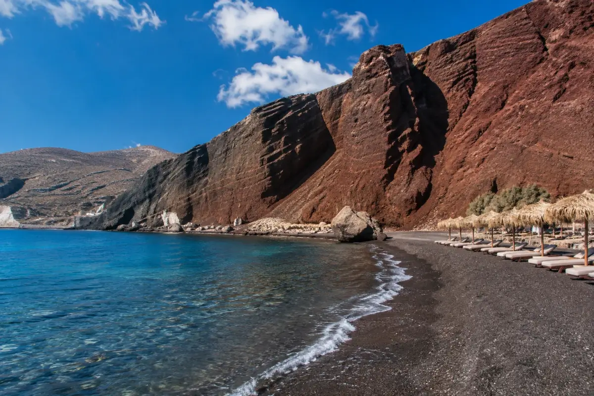 Volcanic beach in Santorini with dark sand, clear water, and dramatic red cliffs