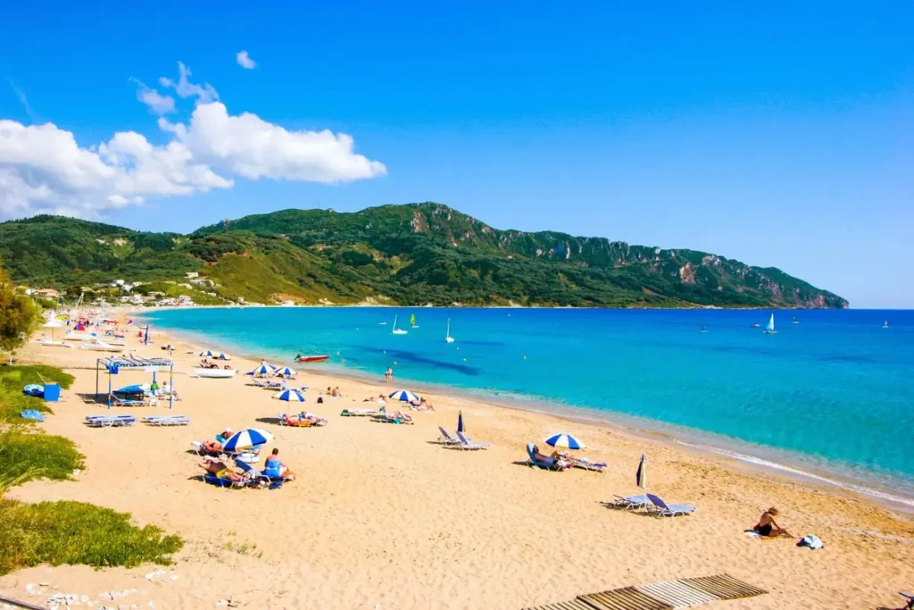 A long sandy beach curves around blue water beneath green hills at Agios Georgios Pagon