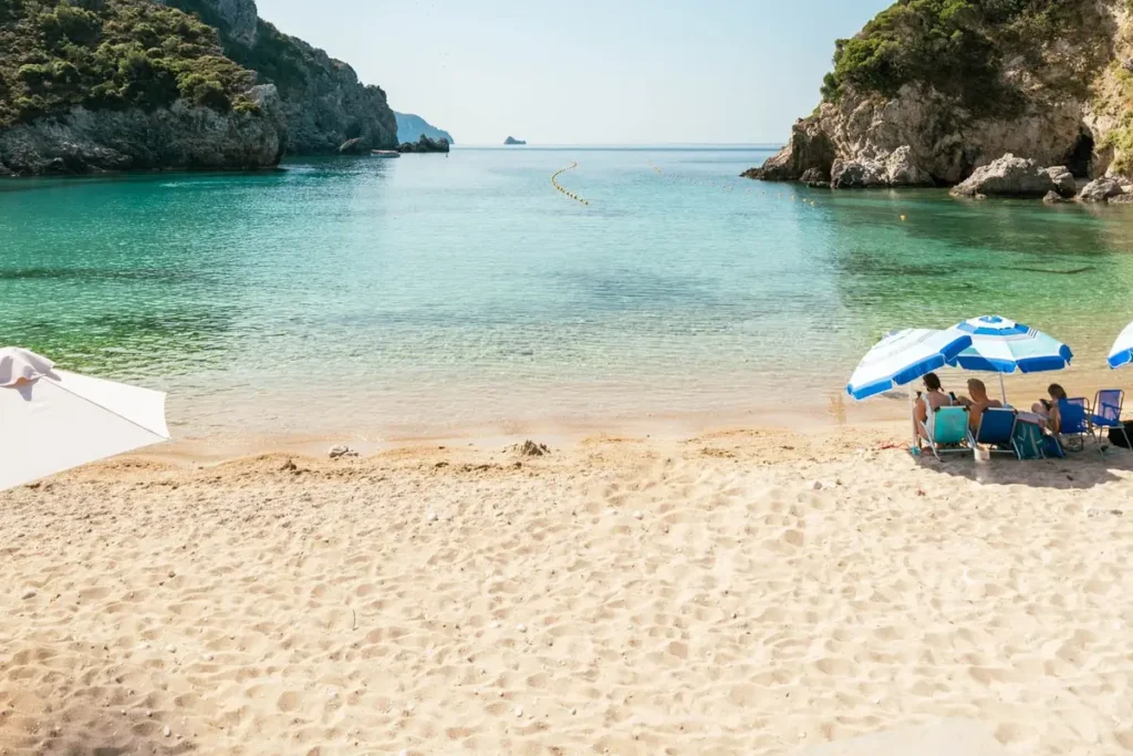 Blue-and-white umbrellas stand on sand beside clear water framed by rocky green headlands