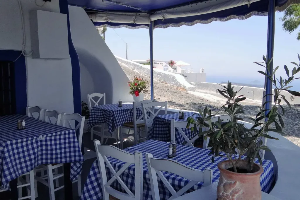 Blue-and-white checked tables and white chairs on Aktaion’s terrace, with the caldera beyond.