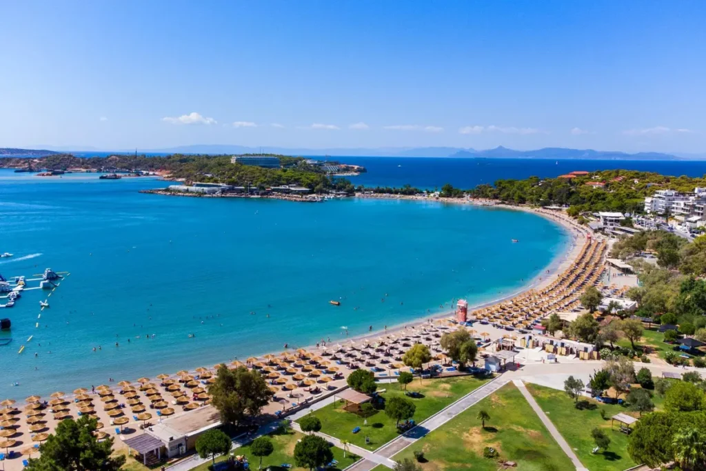 Rows of umbrellas line the curved sandy beach at Akti Vouliagmenis beside blue water.