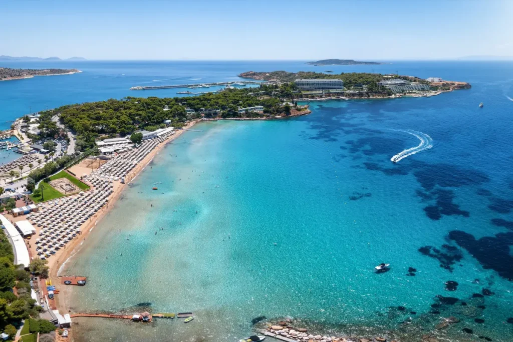 Aerial view of Astir Beach’s umbrella-lined shore and turquoise bay beside a wooded peninsula.