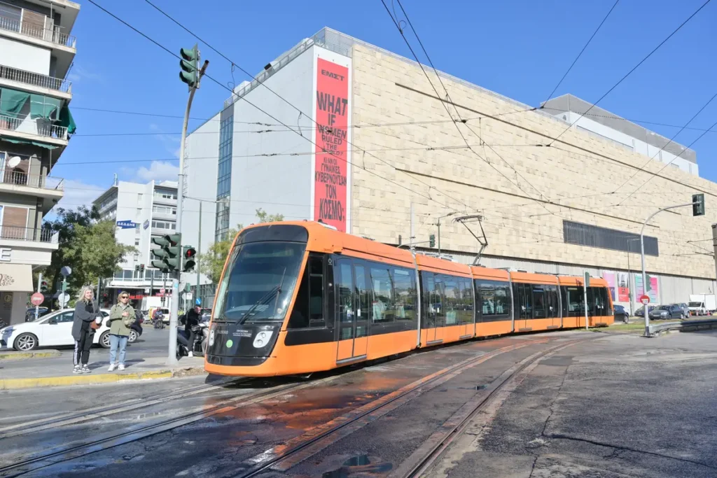 An orange-and-gray Athens tram travels along tracks beside city buildings.