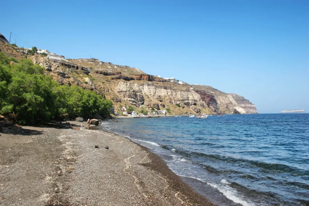 Pebbly shoreline and volcanic cliffs beside the blue water at Caldera Beach in Santorini.