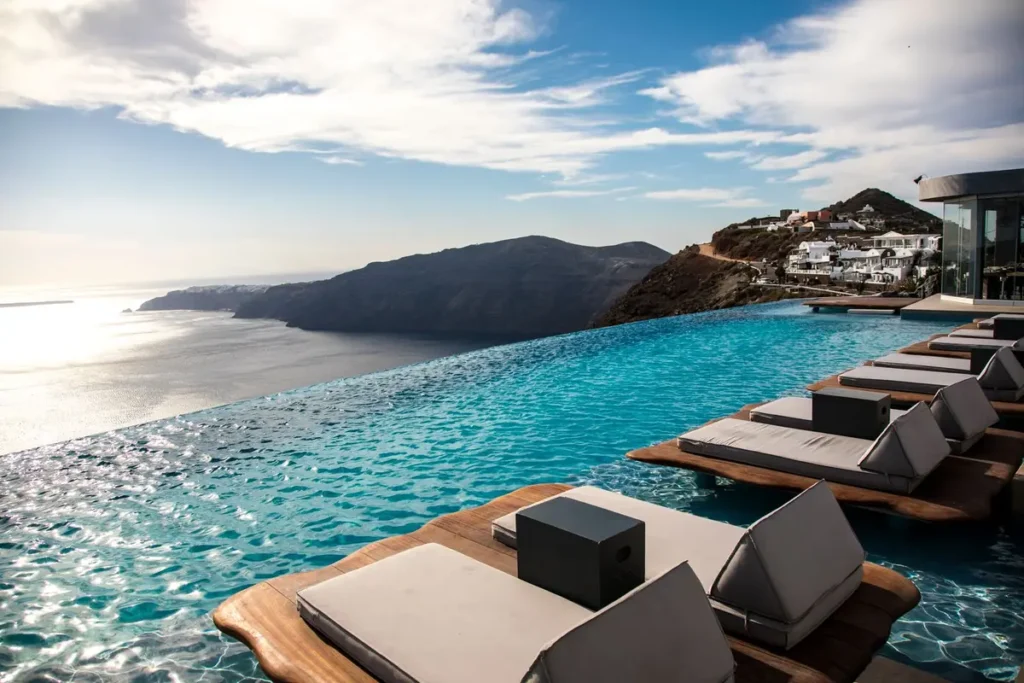 Cushioned loungers on platforms in an infinity pool facing Santorini’s caldera at Cavo Tagoo.