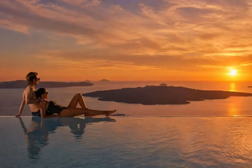 Two guests in an infinity pool overlooking Santorini’s caldera at sunset at Cosmopolitan Suites.