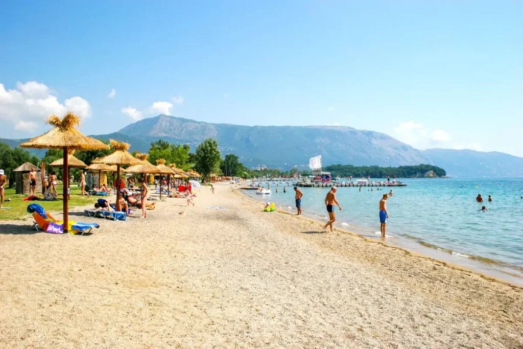 Sun umbrellas and beachgoers along Dassia's shoreline, with green mountains beyond the bay