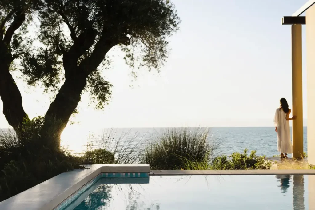 An olive tree shades a pool beside a pale hotel building, with the sea beyond at Domes Miramare