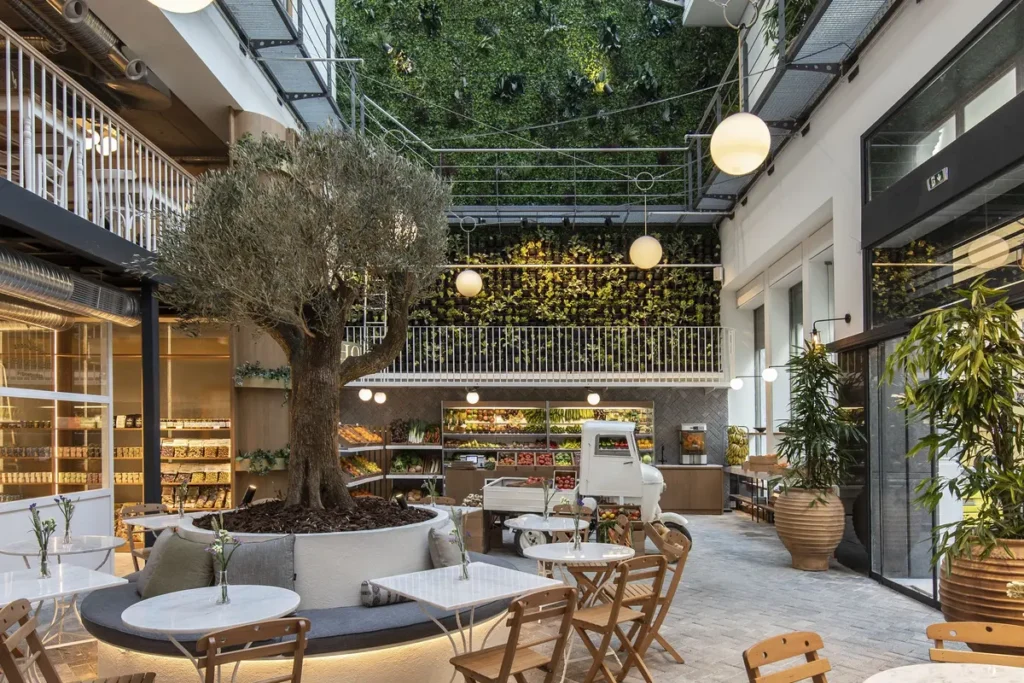 Dining tables and an olive tree beneath a glazed roof and planted wall in Ergon House’s Agora.