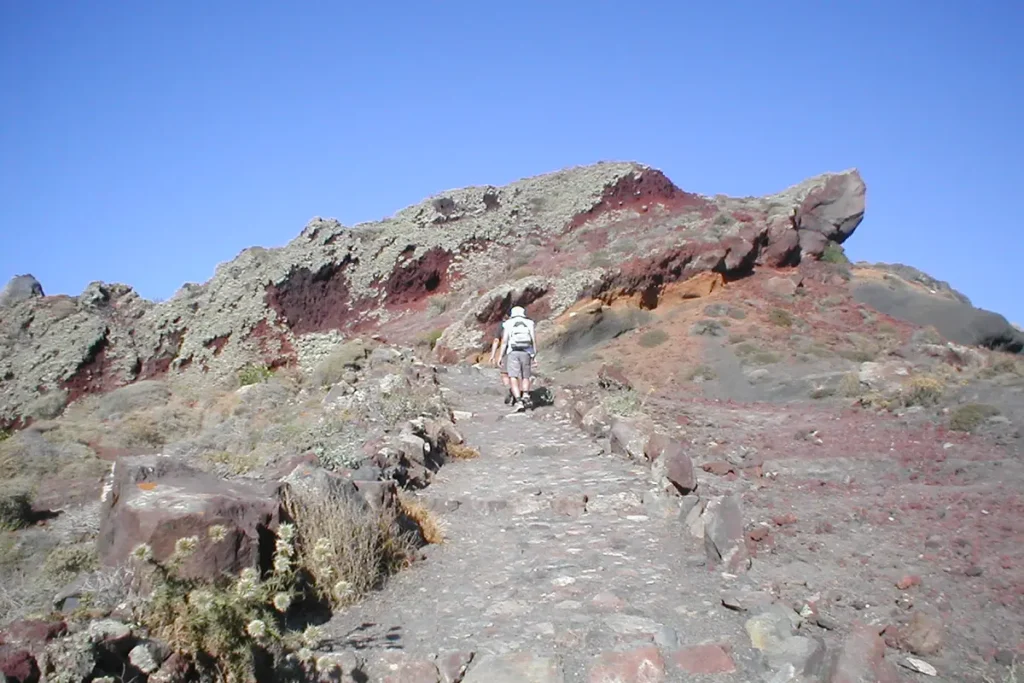 A hiker walking uphill on a rocky path through volcanic terrain between Oia and Fira.