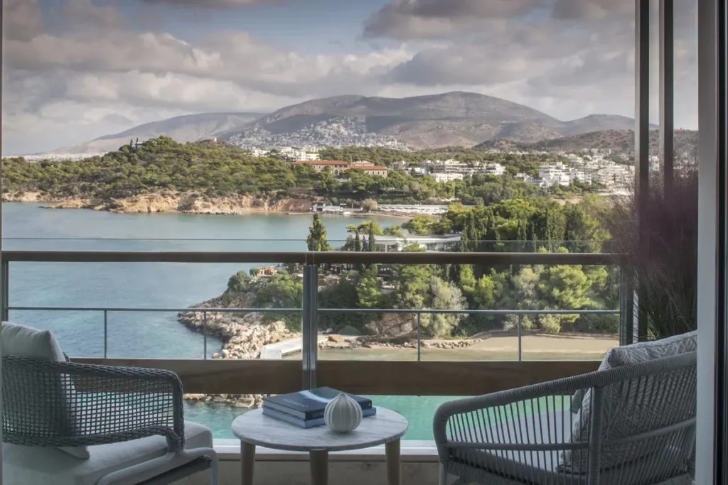 Two chairs and a small table on a Four Seasons Astir Palace balcony overlooking the Vouliagmeni coast.
