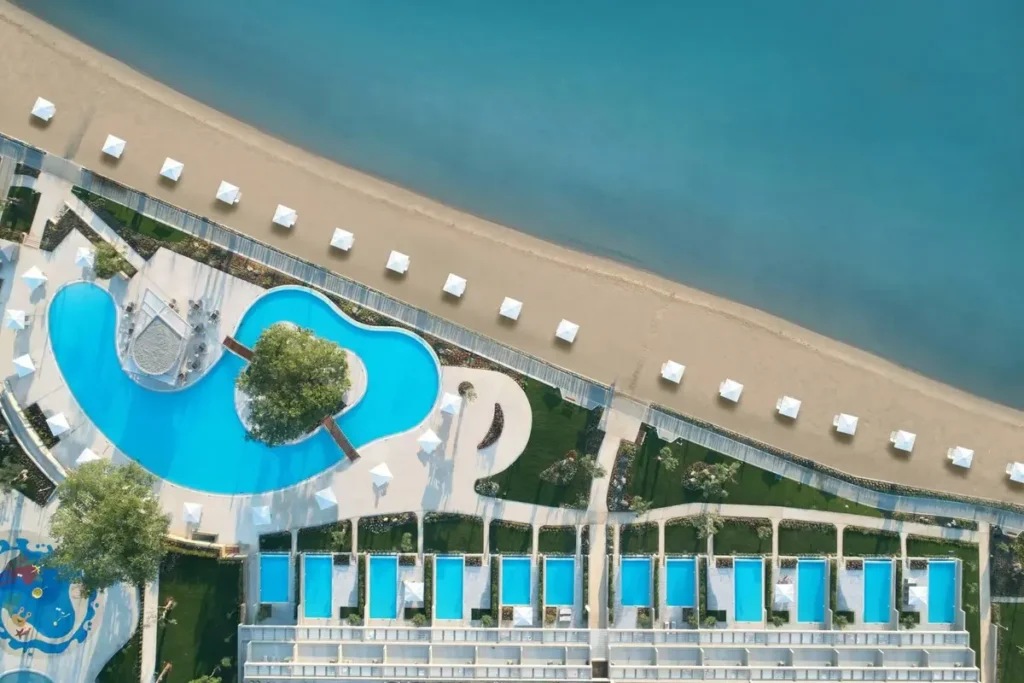 Aerial view of curved swimming pools, landscaped grounds and umbrella rows beside the beach at Ikos Dassia