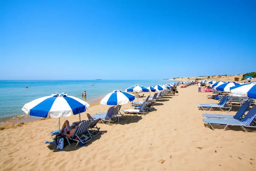 Rows of blue-and-white umbrellas border a long sandy shore and calm blue water at Issos