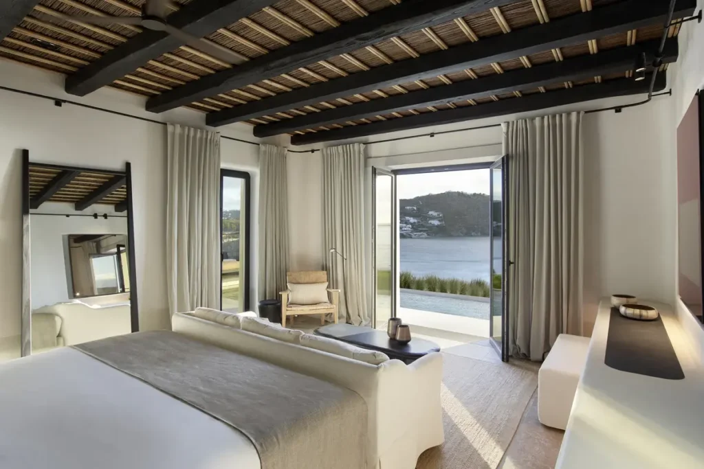 Neutral-toned bedroom with exposed wooden ceiling beams and doors opening toward the sea at Kalesma