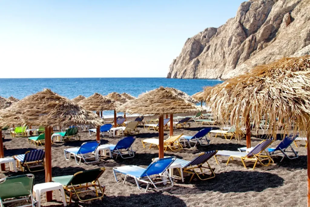 Straw umbrellas and sunbeds on Kamari’s dark shore beneath a rocky headland.