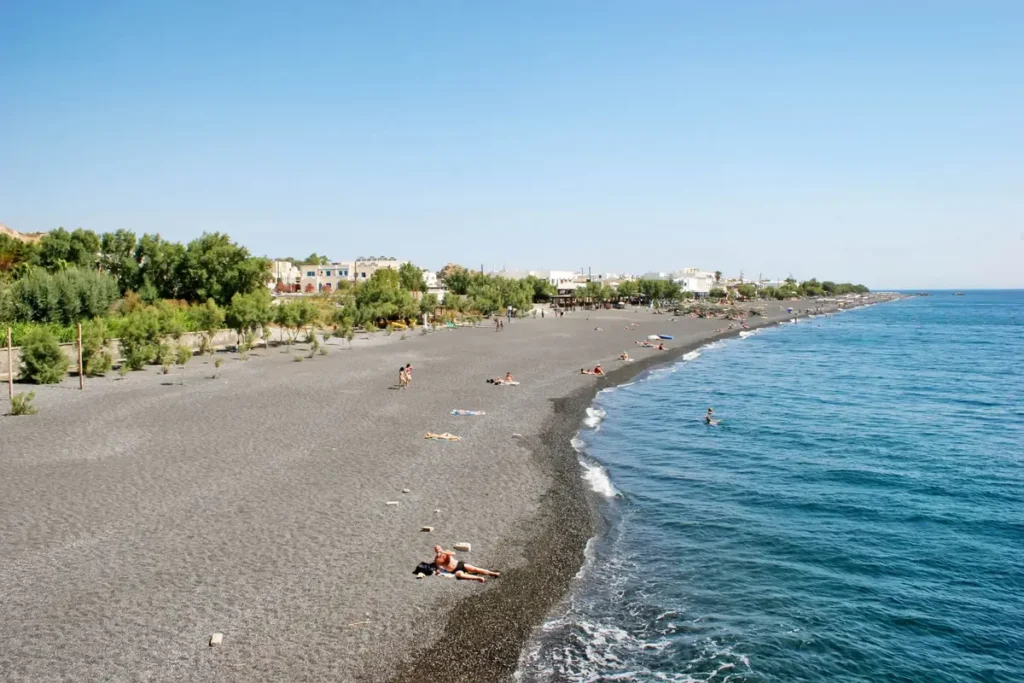 Broad dark volcanic beach beside blue water, with trees and low buildings along Kamari’s coast.