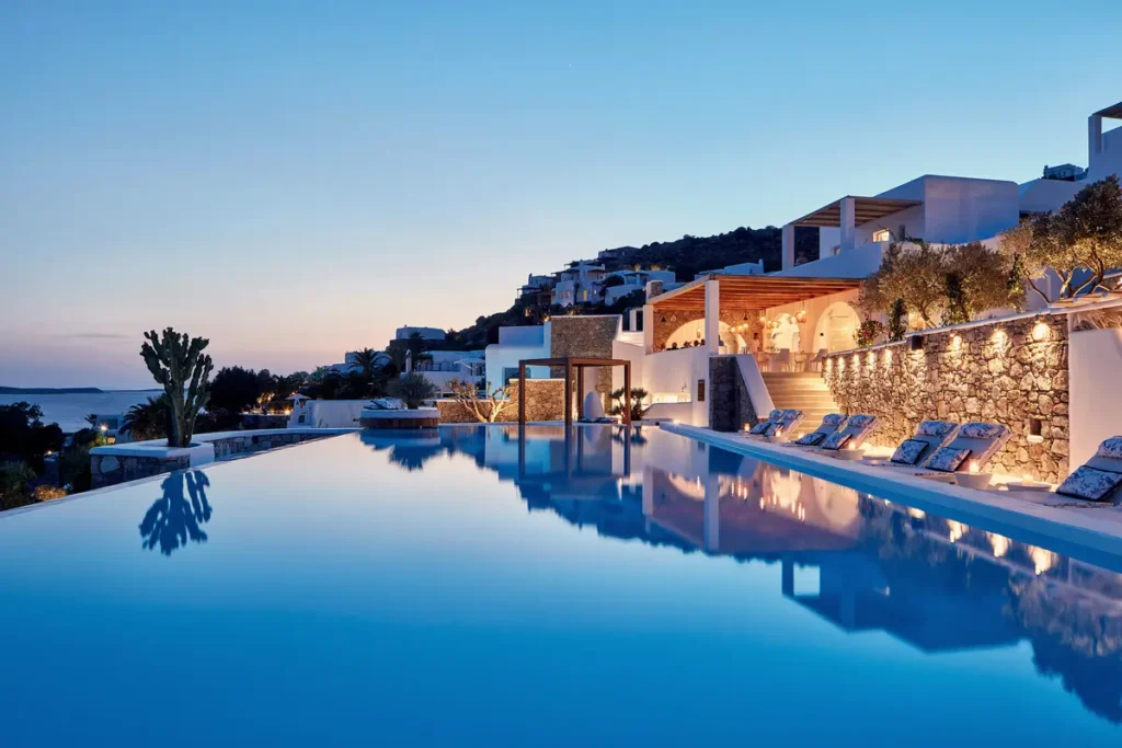 Illuminated pool beside white hillside buildings and stone terraces at Katikies Mykonos at dusk
