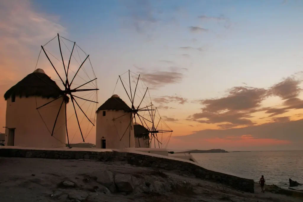 White windmills with dark thatched roofs beside the sea under an orange evening sky in Mykonos
