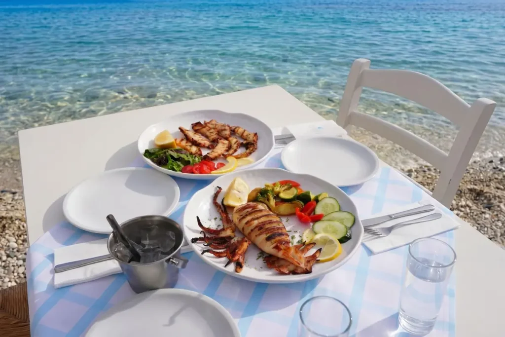 Plates of grilled seafood and vegetables on a white table beside clear seawater at Kavos Taverna