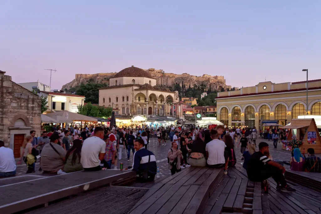 Visitors fill Monastiraki Square as its surrounding buildings light up at dusk.