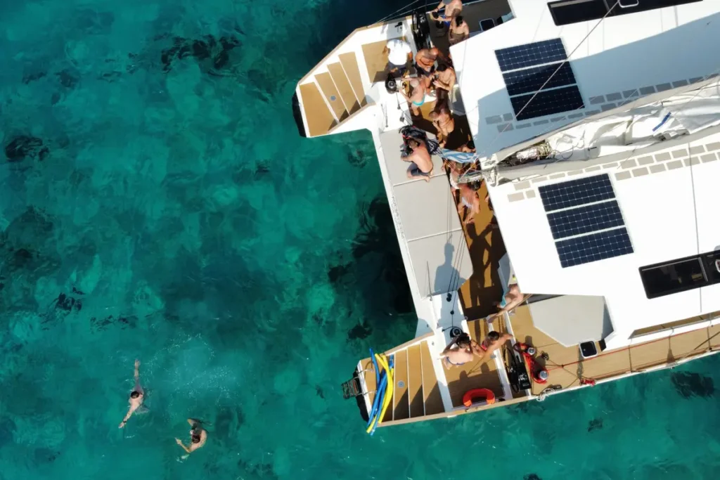 Overhead view of swimmers beside a white catamaran in clear turquoise water near Mykonos