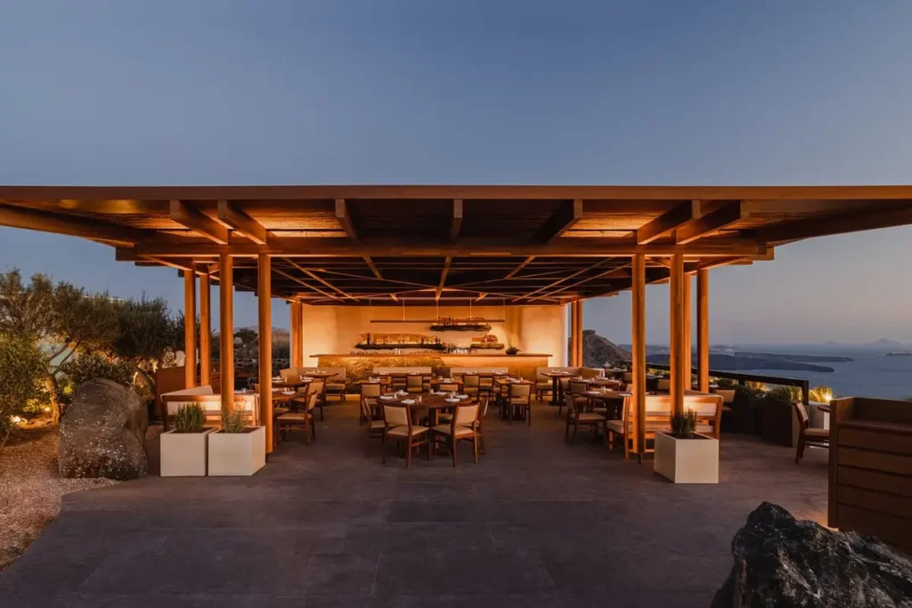 Illuminated timber dining pavilion with tables and caldera views at Nobu Santorini at dusk.
