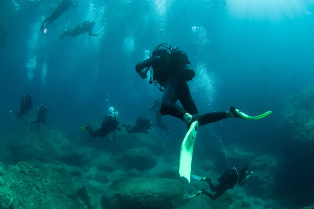 Scuba divers swim above a rocky seabed in blue water with sunlight filtering down