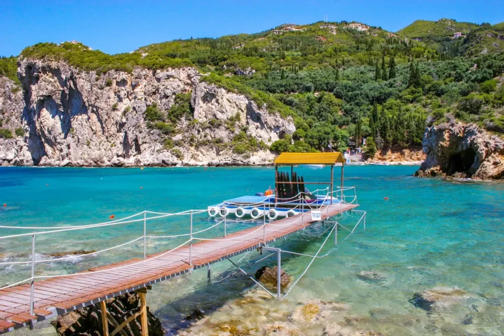 A narrow pontoon extends into turquoise water beneath steep wooded cliffs in Paleokastritsa
