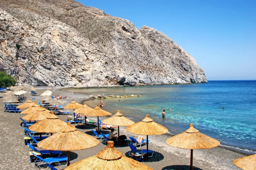 Straw umbrellas and blue loungers beside the dark shore beneath Mesa Vouno at Perissa Beach.