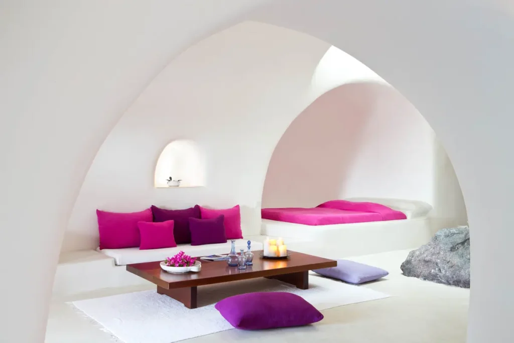 White vaulted suite with magenta cushions, a low wooden table, and a recessed bed at Perivolas in Santorini.