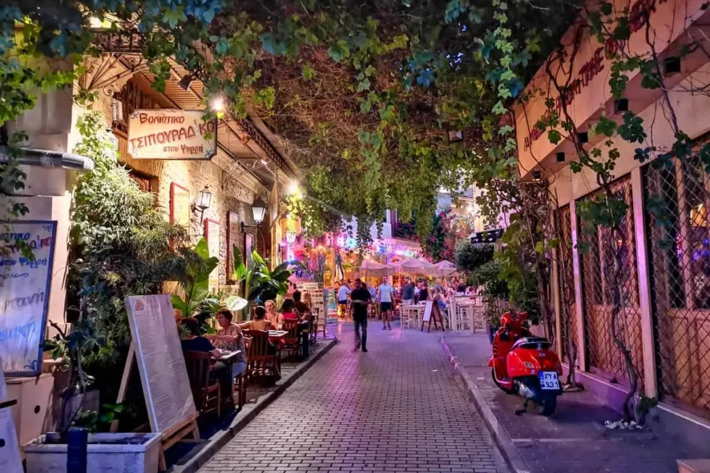 Restaurant tables beneath vines and warm lights along a narrow lane in Psiri at dusk.