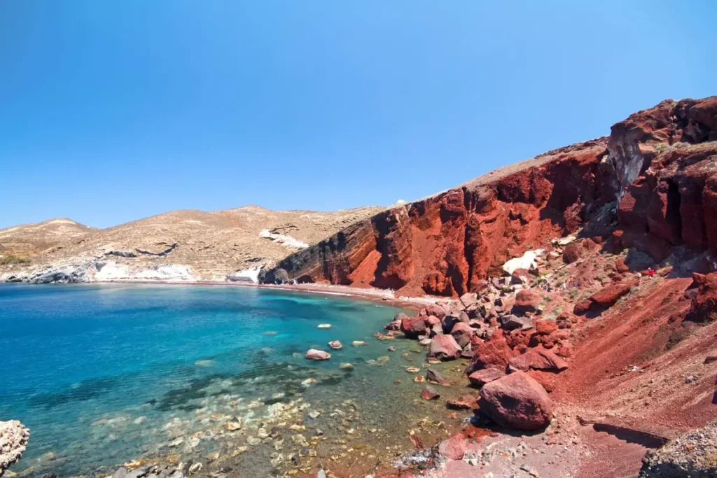 Red volcanic cliffs and dark stones beside clear blue water at Red Beach in Santorini.