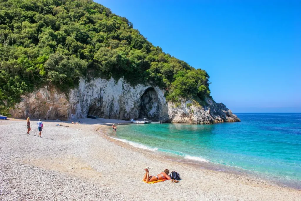A pale beach curves beside clear blue-green water and a wooded rocky headland at Rovinia