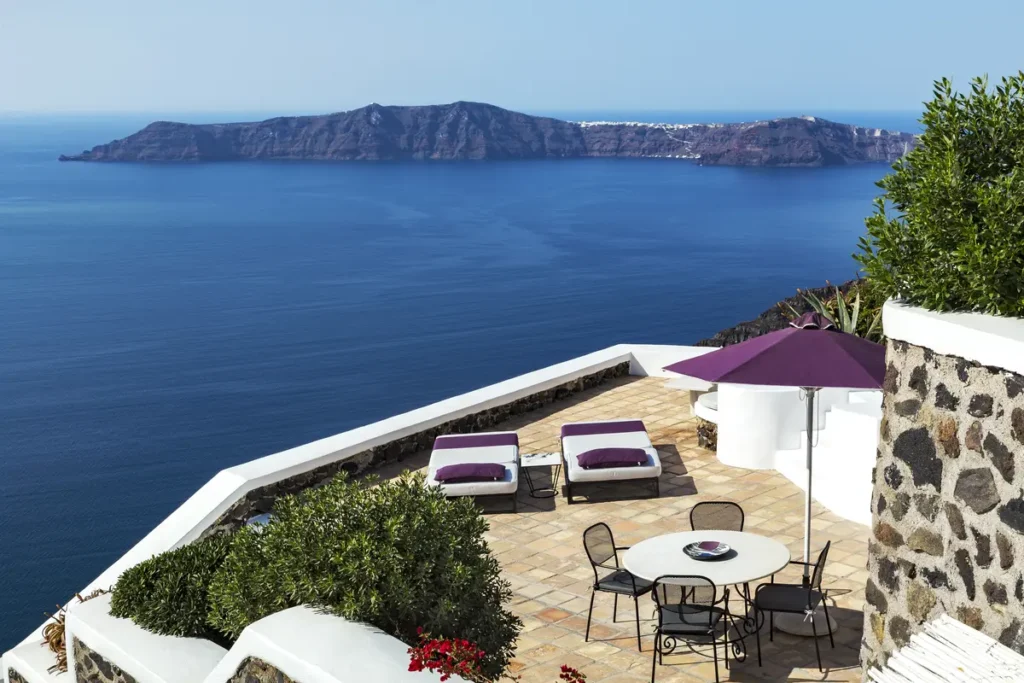 Table, sun loungers and a purple umbrella on a terrace overlooking the caldera at The Vasilicos.