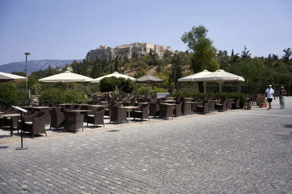 Outdoor café tables beside a broad paved pedestrian route in Thiseio, with the Acropolis beyond.