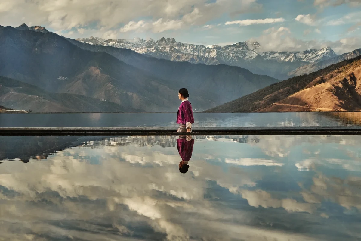 Person beside a reflecting pond mirroring clouds and mountain ridges at Six Senses Bhutan