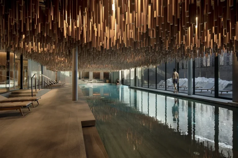 Indoor swimming pool beneath suspended wooden elements with large windows along one side at Six Senses Crans-Montana