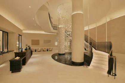 Warm-toned lobby with a curved staircase, tall column, and hanging glass installation at Six Senses London