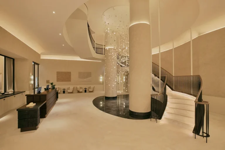 Warm-toned lobby with a curved staircase, tall column, and hanging glass installation at Six Senses London