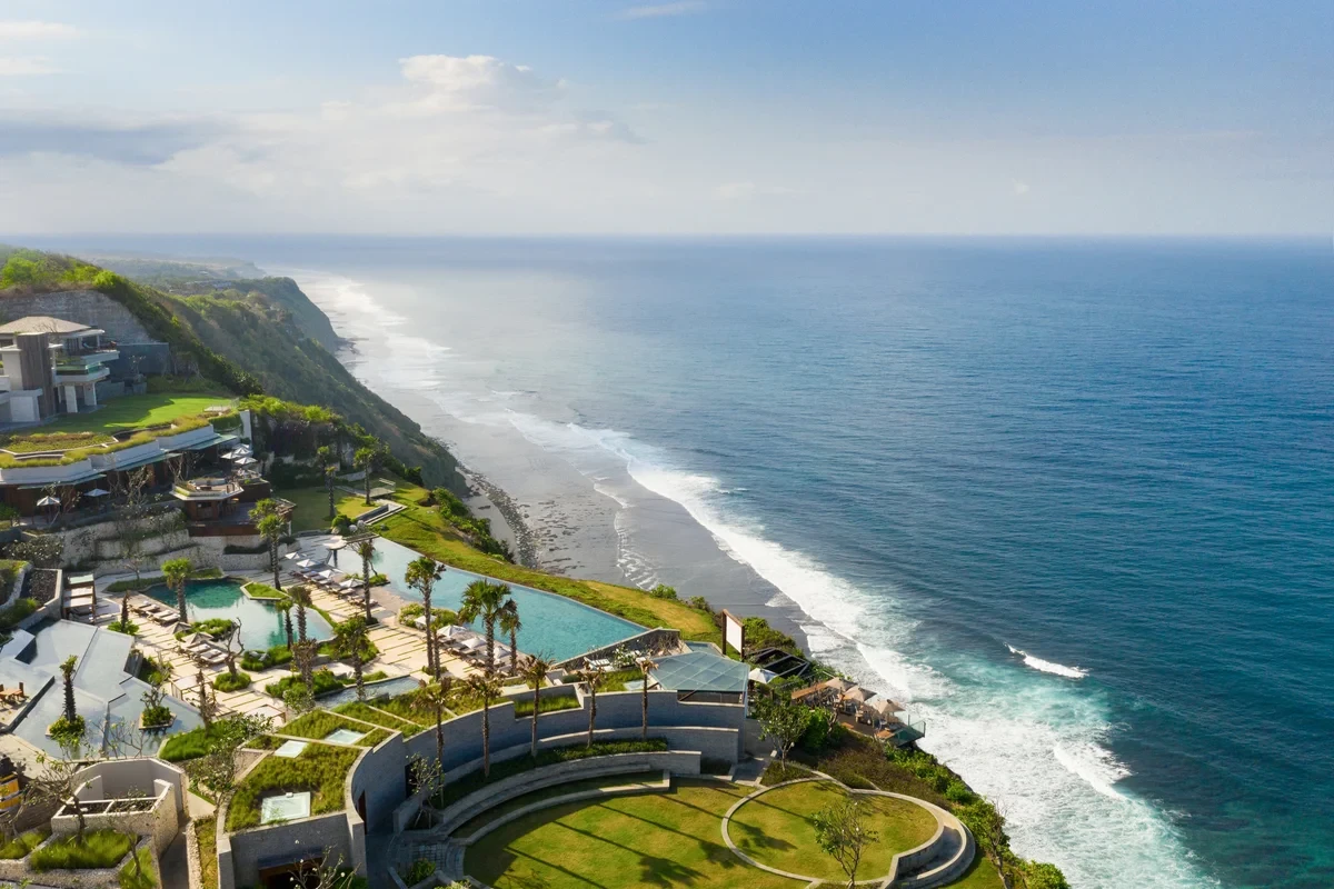 Aerial view of curved resort buildings, pools, and gardens along an ocean cliff at Six Senses Uluwatu Bali