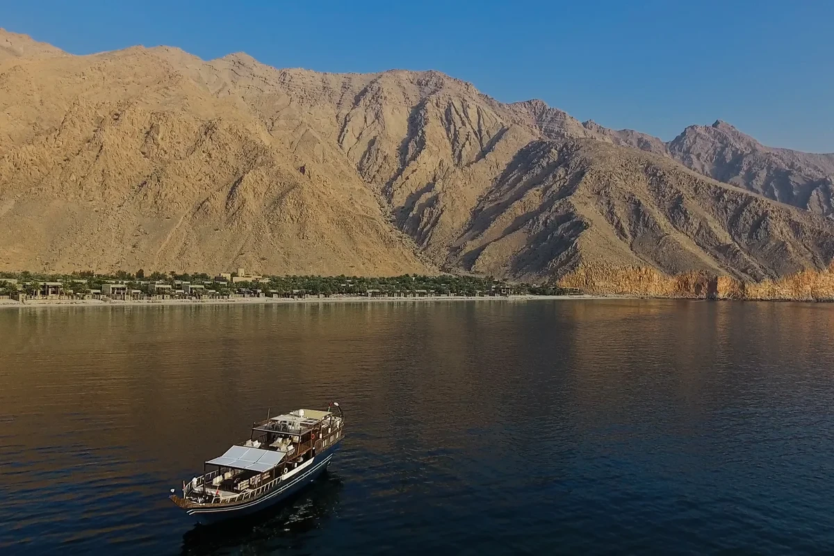 Yacht on calm water below a beach resort and steep, bare mountains at Six Senses Zighy Bay