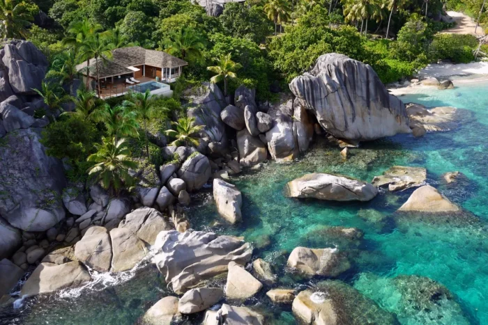 Aerial view of a pool villa among tropical trees and granite boulders beside turquoise water at Six Senses Zil Pasyon
