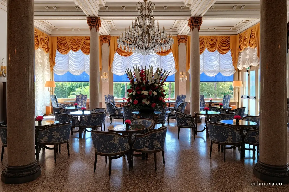 A chandelier hangs above blue upholstered chairs and round tables in an ornate hotel lounge.