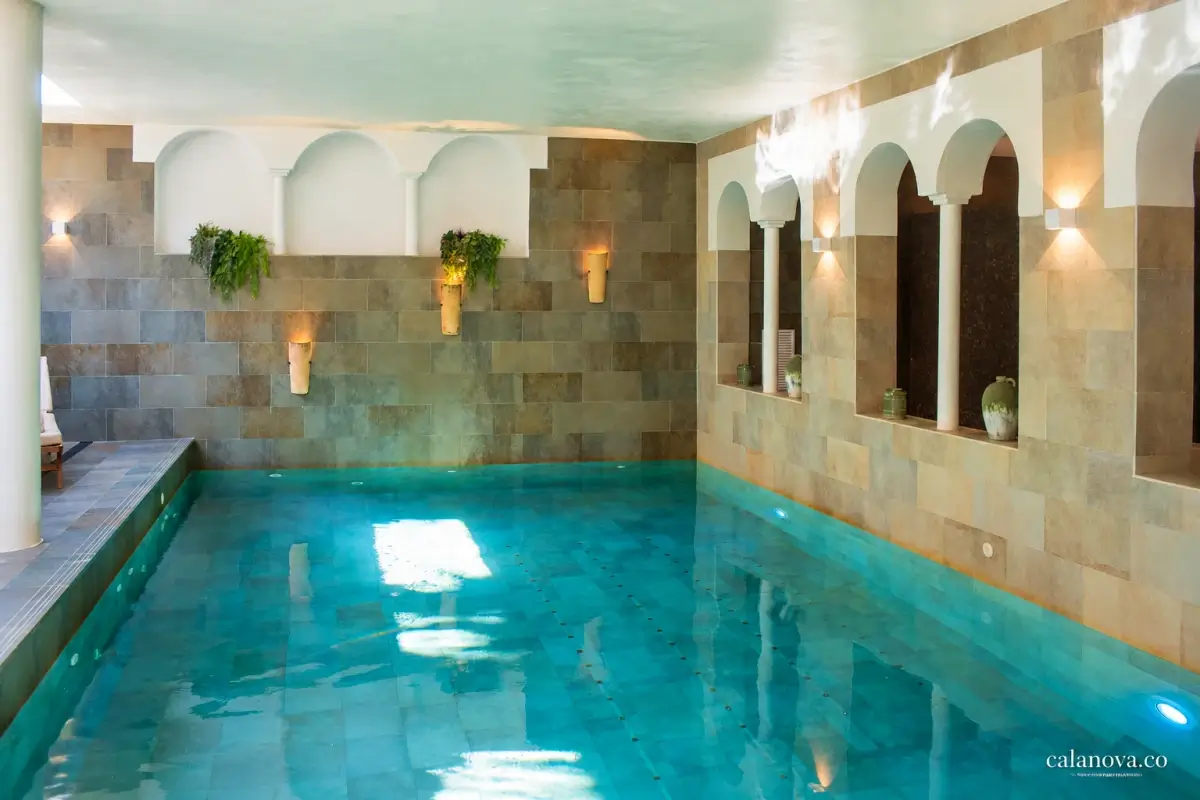 A turquoise indoor pool beneath reflected daylight, with stone walls, arched alcoves and hanging plants.