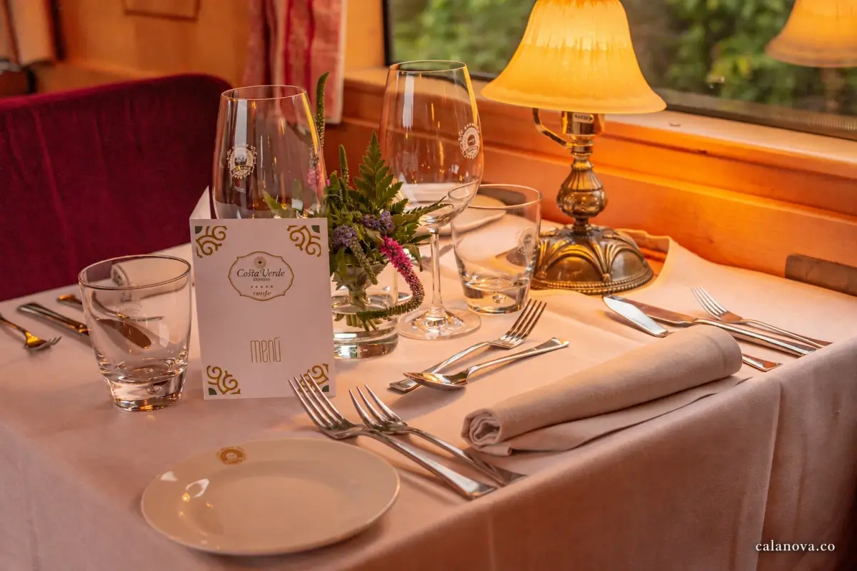 A train dining table set with white linen, glassware, silver cutlery and a brass lamp beside the window.