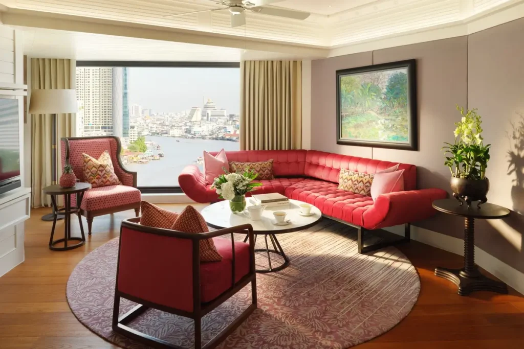 Red sofa and armchairs around a coffee table beside a river-facing window in the Premier One-Bedroom Suite at Mandarin Oriental, Bangkok.