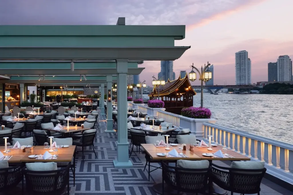 Outdoor dining tables, a moored boat, and the Chao Phraya skyline at dusk at Riverside Terrace, Mandarin Oriental, Bangkok.