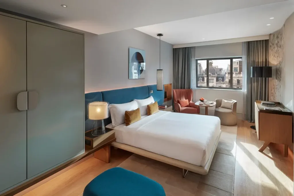 Bed with a blue headboard, seating beside the window, and neighboring architecture visible from a Deluxe Boulevard Room at Mandarin Oriental, Barcelona.