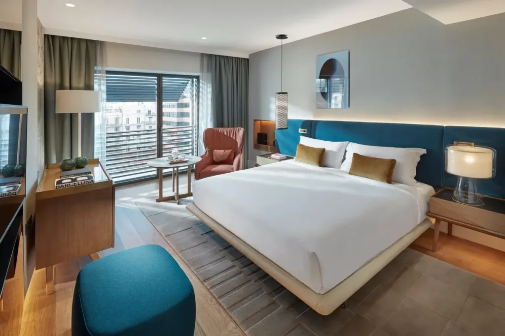 Bed with a blue headboard, seating, and a window with exterior screening in a Deluxe Garden Room at Mandarin Oriental, Barcelona.