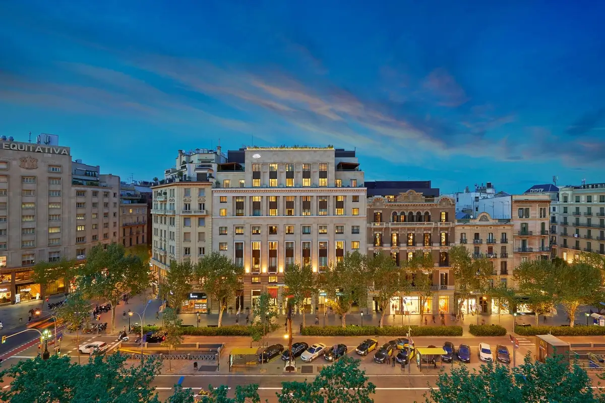 Illuminated facade of Mandarin Oriental, Barcelona facing Passeig de Gràcia, with traffic, trees, and neighboring buildings at dusk.